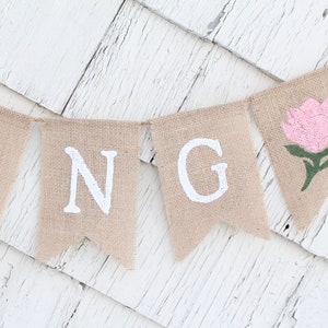 May include: A burlap banner with the word "SPRING" spelled out in white paint. The "G" is followed by a pink flower with green leaves.