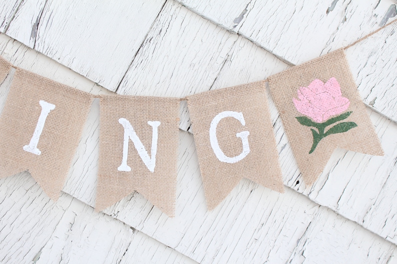 May include: A burlap banner with the word "SPRING" spelled out in white paint. The "G" is followed by a pink flower with green leaves.