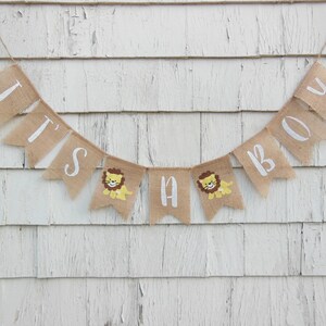 May include: A burlap banner with the words "It's a Boy" in white paint. The banner has small lion illustrations on each of the flags.
