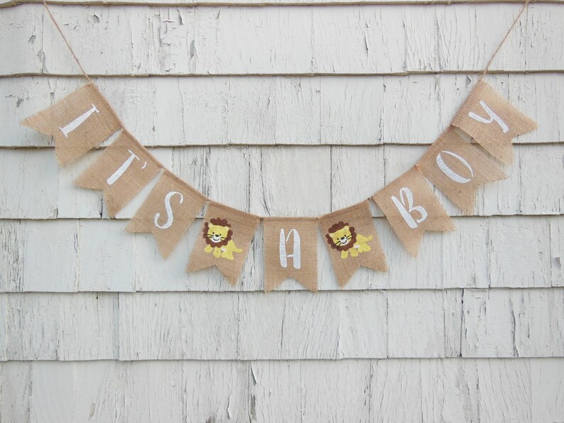 May include: A burlap banner with the words "It's a Boy" in white paint. The banner has small lion illustrations on each of the flags.