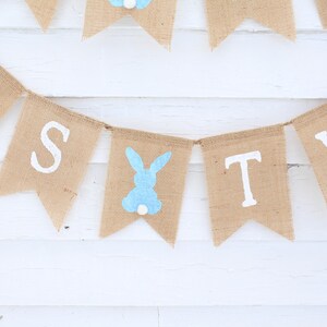 May include: A burlap banner with the words "It's Two" in white paint. A blue bunny silhouette is between the words "Two" and "It's".