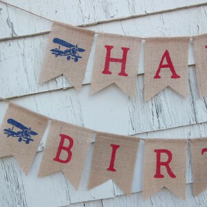 Może przedstawiać: Baner z juty z napisem "Happy Birthday" w czerwonych literach. Każda litera znajduje się na osobnej fladze. Flagi są ozdobione niebieskimi samolotami.