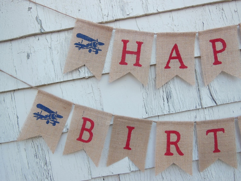 Może przedstawiać: Baner z juty z napisem "Happy Birthday" w czerwonych literach. Każda litera znajduje się na osobnej fladze. Flagi są ozdobione niebieskimi samolotami.