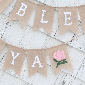 Puede incluir: Una pancarta de arpillera con las palabras "Bless Ya" pintadas en blanco. La pancarta tambi&eacute;n presenta una flor rosa con hojas verdes.