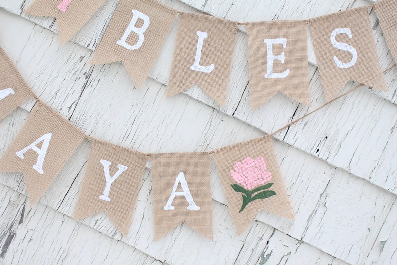 Puede incluir: Una pancarta de arpillera con las palabras "Bless Ya" pintadas en blanco. La pancarta tambi&eacute;n presenta una flor rosa con hojas verdes.