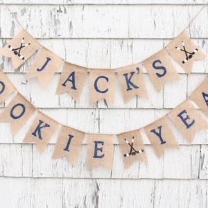 Pu&ograve; includere: Uno striscione di juta con il testo "JACK'S ROOKIE YEAR" in lettere blu scuro. Lo striscione &egrave; decorato con bastoni da hockey e dischi.