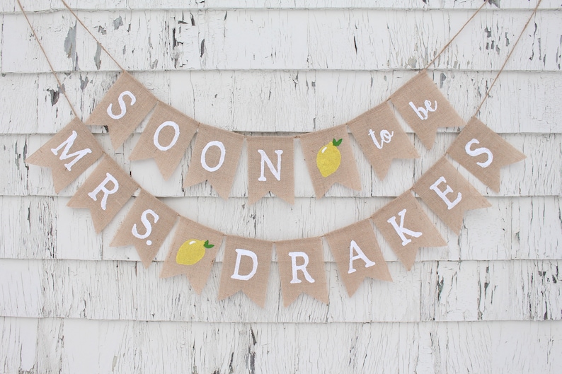 May include: A burlap banner with the text "Mrs. Soon to be Drakes" in white letters. The banner has small yellow lemons with green stems on each flag.