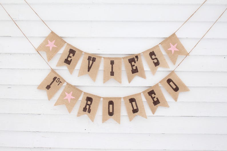 Pode incluir: Uma faixa de serapilheira com o texto "EVIE'S 1st RODEO" em letras castanhas escuras. Acentos de estrelas cor-de-rosa est&atilde;o em algumas das bandeirinhas. A faixa &eacute; amarrada em barbante e pendurada em um fundo de madeira branca.