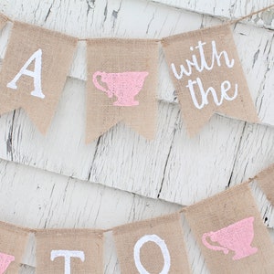 May include: A burlap banner with the words "Tea with the" and a pink teacup graphic. The banner is strung with white twine and hangs against a white wood background.