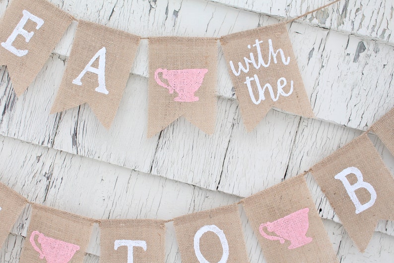 May include: A burlap banner with the words "Tea with the" and a pink teacup graphic. The banner is strung with white twine and hangs against a white wood background.