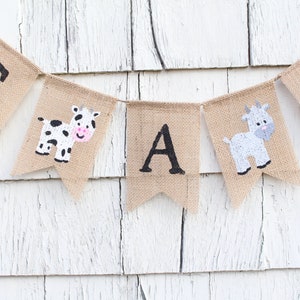Puede incluir: Una pancarta de arpillera con las letras "It's a Boy" en pintura negra. La pancarta est&aacute; decorada con una vaca y una cabra.