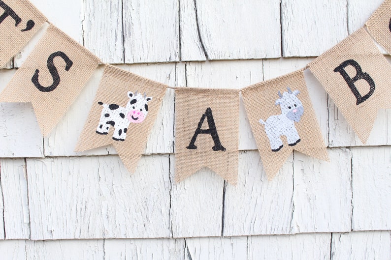 Puede incluir: Una pancarta de arpillera con las letras "It's a Boy" en pintura negra. La pancarta est&aacute; decorada con una vaca y una cabra.