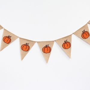 May include: A bunting garland made of hessian fabric, with triangular flags. Each flag displays an orange pumpkin design with a brown stalk. The garland is strung on twine, ideal for autumn or fall decorations.