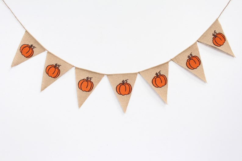 May include: A bunting garland made of hessian fabric, with triangular flags. Each flag displays an orange pumpkin design with a brown stalk. The garland is strung on twine, ideal for autumn or fall decorations.