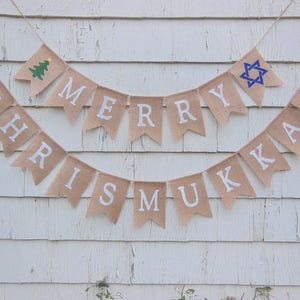 May include: Burlap banner with the words "MERRY CHRISTMUKKAH" in white letters. The banner includes a green Christmas tree and a blue Star of David. The banner is hanging against a white wooden background.