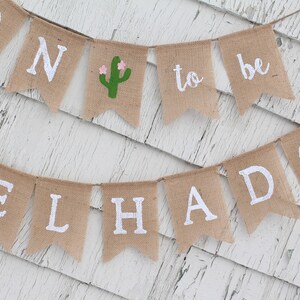 May include: A burlap banner with the words "Soon to be Melhado" in white lettering. The banner is decorated with a green cactus with pink flowers.