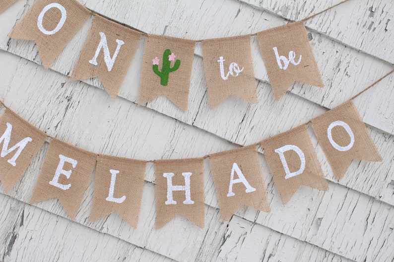 May include: A burlap banner with the words "Soon to be Melhado" in white lettering. The banner is decorated with a green cactus with pink flowers.
