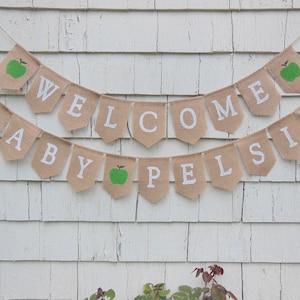 K&ouml;nnte beinhalten: Ein Wimpelbanner aus Jute mit den Worten "WELCOME BABY PELSIS" in wei&szlig;en Buchstaben. Einige Wimpel zeigen gr&uuml;ne &Auml;pfel. Der Banner h&auml;ngt vor einem verwitterten, wei&szlig;en Holzuntergrund.