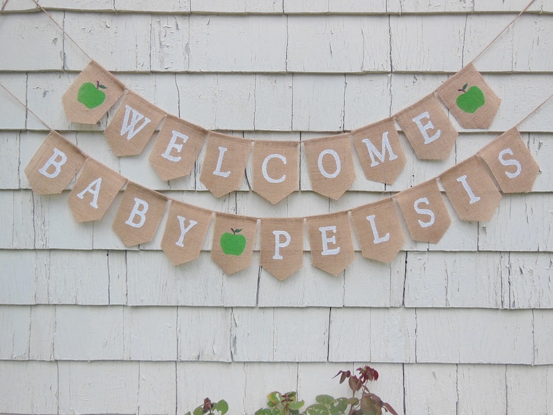 K&ouml;nnte beinhalten: Ein Wimpelbanner aus Jute mit den Worten "WELCOME BABY PELSIS" in wei&szlig;en Buchstaben. Einige Wimpel zeigen gr&uuml;ne &Auml;pfel. Der Banner h&auml;ngt vor einem verwitterten, wei&szlig;en Holzuntergrund.