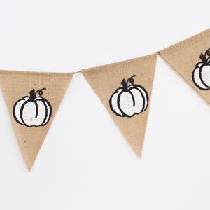 May include: A hessian banner featuring triangular flags, each printed with a white pumpkin design with black outlines and stems. The banner is strung on twine, ideal for autumn decorations.