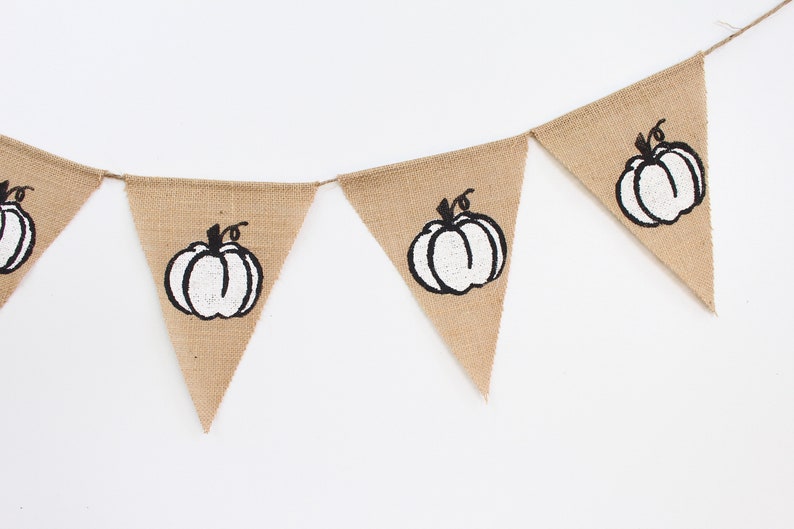 May include: A hessian banner featuring triangular flags, each printed with a white pumpkin design with black outlines and stems. The banner is strung on twine, ideal for autumn decorations.