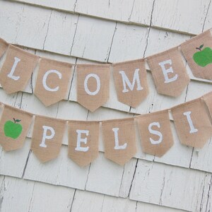 K&ouml;nnte beinhalten: Ein Wimpelbanner aus Jute mit wei&szlig;en Buchstaben, die "WELCOME PELSIS" bilden. Das Banner enth&auml;lt zwei gr&uuml;ne Apfelgrafiken. Es h&auml;ngt vor einem wei&szlig;en, verwitterten Holzhintergrund.