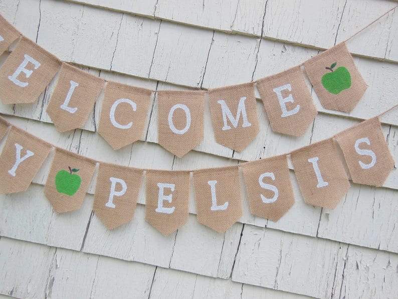 K&ouml;nnte beinhalten: Ein Wimpelbanner aus Jute mit wei&szlig;en Buchstaben, die "WELCOME PELSIS" bilden. Das Banner enth&auml;lt zwei gr&uuml;ne Apfelgrafiken. Es h&auml;ngt vor einem wei&szlig;en, verwitterten Holzhintergrund.