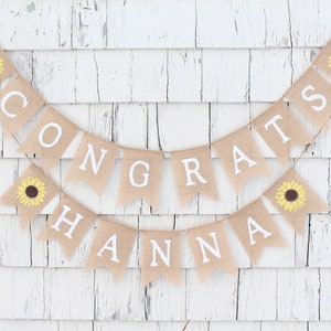 May include: A burlap banner with the words "CONGRATS HANNA" in white letters. The banner is decorated with yellow sunflowers and hangs against a white, weathered wood background. A festive decoration for a celebration.