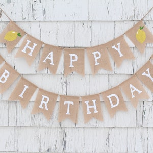 May include: A burlap banner with the words "Happy Birthday" in white letters and lemon illustrations. The banner is strung on a white wooden wall.