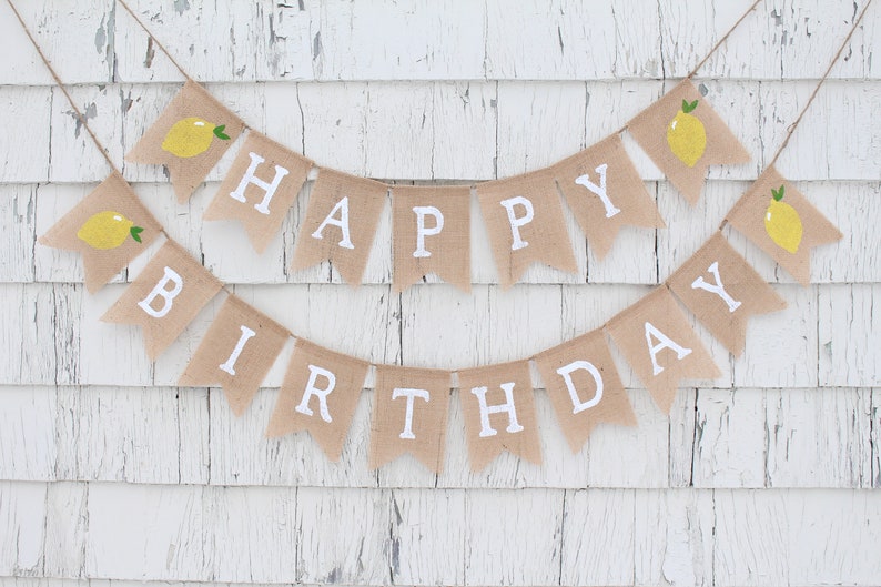 May include: A burlap banner with the words "Happy Birthday" in white letters and lemon illustrations. The banner is strung on a white wooden wall.