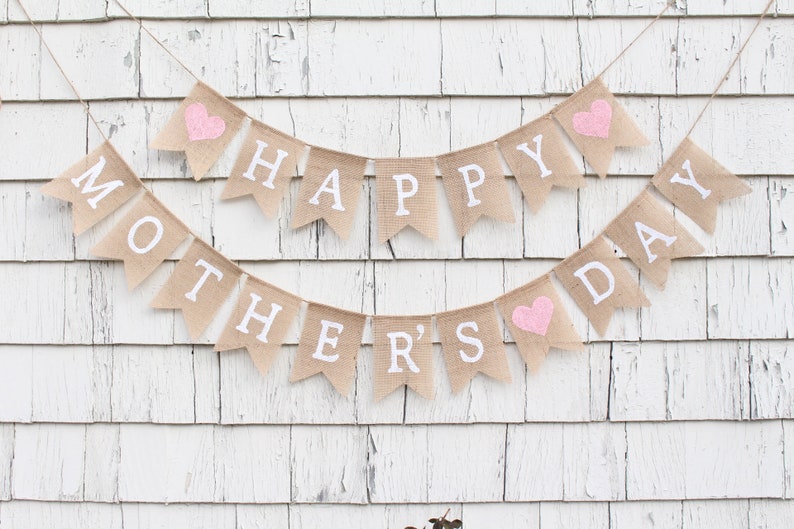 May include: A burlap banner with the words "Happy Mother's Day" in white letters. The banner has pink hearts on each end.