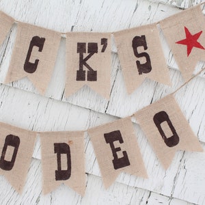 Pode incluir: Uma faixa de serapilheira com as palavras "JACK'S RODEO" impressas em letras escuras. Uma estrela vermelha adorna a extremidade direita. A faixa est&aacute; pendurada em uma corda, em um fundo de madeira branca desgastada.