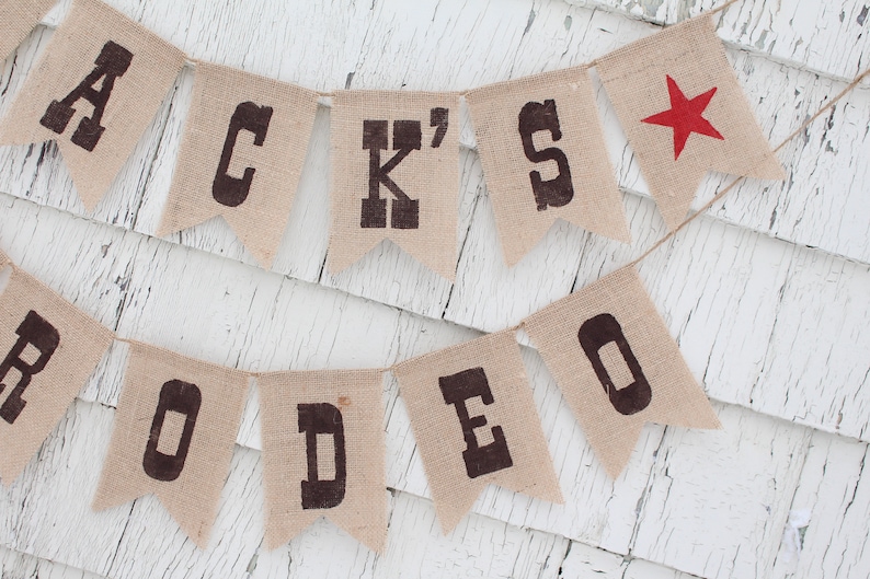 Pode incluir: Uma faixa de serapilheira com as palavras "JACK'S RODEO" impressas em letras escuras. Uma estrela vermelha adorna a extremidade direita. A faixa est&aacute; pendurada em uma corda, em um fundo de madeira branca desgastada.