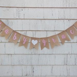 May include: A burlap banner with pink lettering that reads "baby grace" with a white heart in between the words.