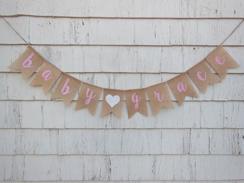 May include: A burlap banner with pink lettering that reads "baby grace" with a white heart in between the words.
