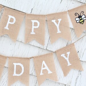 May include: A burlap banner with the words "Happy Day" in white paint. The banner is decorated with two yellow and black bees.