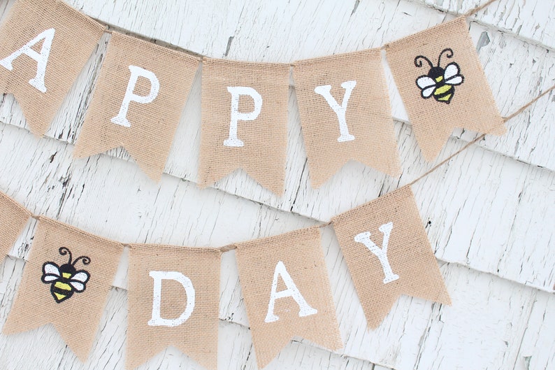 May include: A burlap banner with the words "Happy Day" in white paint. The banner is decorated with two yellow and black bees.