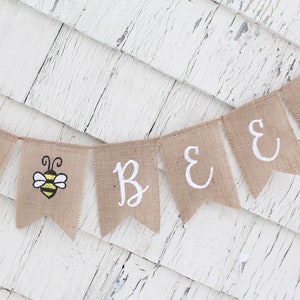 May include: A burlap banner with the words "Bee" and a bee illustration. The banner is strung on a white wooden background.