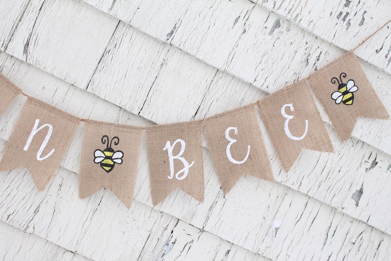 May include: A burlap banner with the words "Bee" and a bee illustration. The banner is strung on a white wooden background.