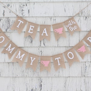 May include: A burlap banner with the text "Mommy to Be" and "Tea with the" with pink teacups on each side. The banner is hanging on a white wooden wall.