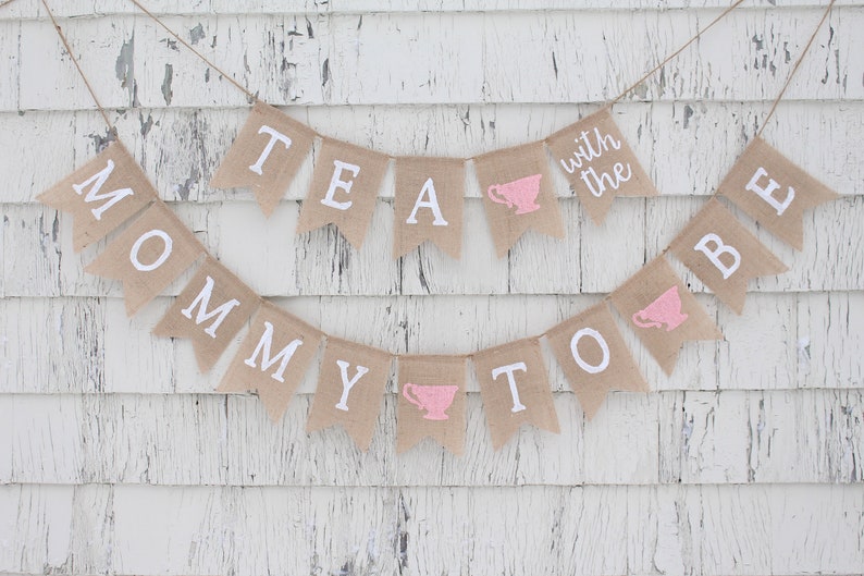 May include: A burlap banner with the text "Mommy to Be" and "Tea with the" with pink teacups on each side. The banner is hanging on a white wooden wall.