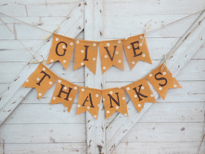 May include: A festive banner with the words "GIVE THANKS" in dark brown letters. The pennants are a light orange color with white polka dots, strung on twine, and set against a white wooden background.