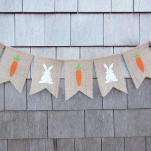 May include: A burlap banner with alternating images of white bunnies and orange carrots. The banner is strung on a wall with a wood grain pattern.