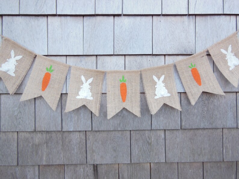 May include: A burlap banner with alternating images of white bunnies and orange carrots. The banner is strung on a wall with a wood grain pattern.