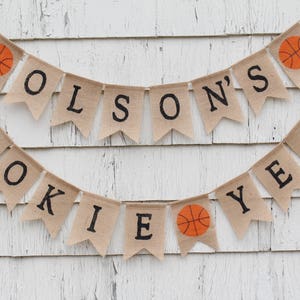 Pu&ograve; includere: Uno striscione di juta con il testo "ROOKIE YEAR" e "OLSON'S" con palloni da basket tra le parole. Lo striscione &egrave; appeso a un muro di legno bianco.
