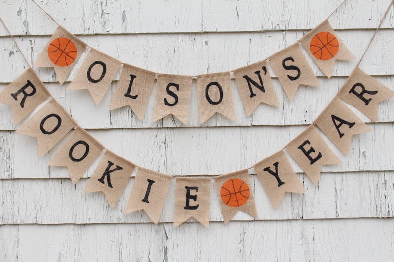 Pu&ograve; includere: Uno striscione di juta con il testo "ROOKIE YEAR" e "OLSON'S" con palloni da basket tra le parole. Lo striscione &egrave; appeso a un muro di legno bianco.