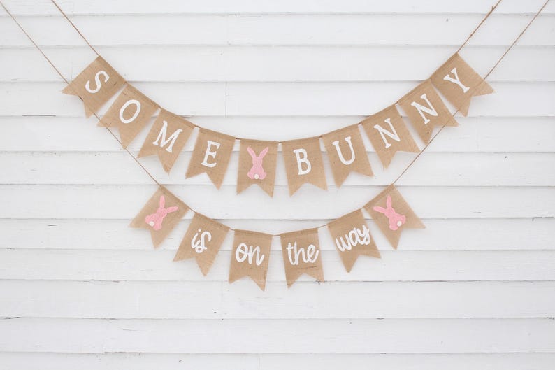 May include: A burlap banner with pink bunny cutouts and the text "Some bunny is on the way".