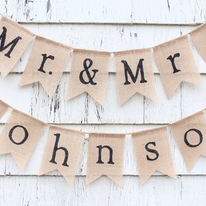 K&ouml;nnte beinhalten: Ein Banner aus Jute mit dem Text "Mr. & Mrs. Johnson" in schwarzen Buchstaben. Das Banner ist an einer wei&szlig;en Holzwand befestigt.