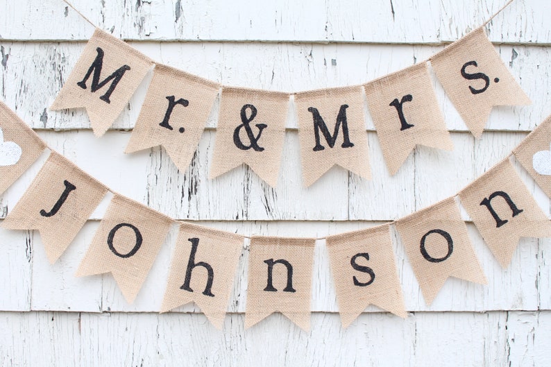 K&ouml;nnte beinhalten: Ein Banner aus Jute mit dem Text "Mr. & Mrs. Johnson" in schwarzen Buchstaben. Das Banner ist an einer wei&szlig;en Holzwand befestigt.