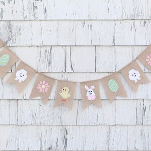 K&ouml;nnte beinhalten: Ein Osterbanner aus Jute mit einer Reihe von Flaggen, die verschiedene Ostermotive zeigen, darunter ein H&auml;schen, ein K&uuml;ken, ein Ei, eine Blume und ein Lamm. Das Banner h&auml;ngt vor einem wei&szlig;en Holz Hintergrund.